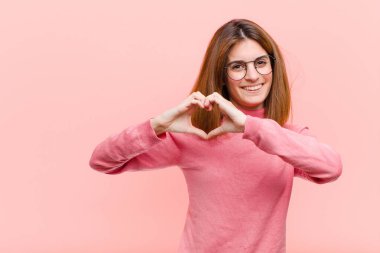 Genç ve güzel bir kadın gülümsüyor ve mutlu, sevimli, romantik ve aşık, pembe arka planda iki eliyle kalp şekli oluşturuyor.
