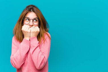 Genç, güzel bir kadın endişeli, gergin, stresli ve korkmuş görünüyor. Tırnaklarını yiyor ve mavi arka plana karşı yanal fotokopi alanı arıyor.