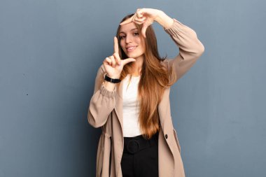 Genç, güzel bir kadın mutlu, arkadaş canlısı ve pozitif hissediyor, gülümsüyor ve elleri gri arka planda bir portre veya fotoğraf çerçevesi yapıyor.