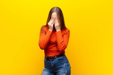 young pretty woman feeling sad, frustrated, nervous and depressed, covering face with both hands, crying against orange background