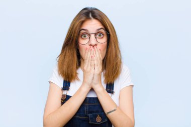 Genç, güzel bir kadın endişeli, üzgün ve korkmuş hissediyor. Elleriyle ağzını kapatıyor. Endişeli görünüyor ve mavi arka planı mahvediyor.