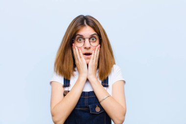 Genç ve güzel bir kadın şok olmuş ve korkmuş hissediyor. Ağzı açık ve elleri mavi arka planda.