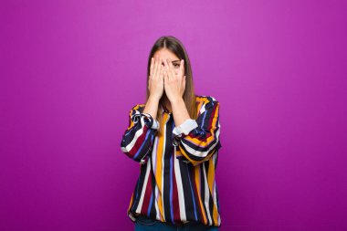 Genç ve güzel bir kadın elleriyle yüzünü kapatıyor, parmaklarının arasından şaşkın bir ifadeyle bakıyor ve mor arka plana bakıyor.