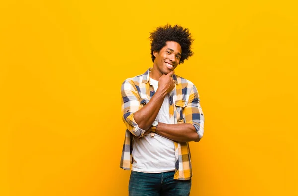 young black man smiling, enjoying life, feeling happy, friendly, satisfied and carefree with hand on chin against orange wall