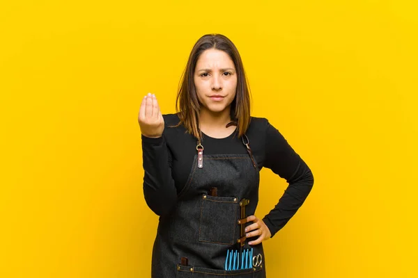 hairdresser woman making capice or money gesture, telling you to pay your debts! against orange background