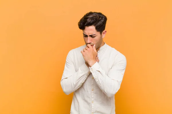 young handsome man feeling ill with a sore throat and flu symptoms, coughing with mouth covered against orange background