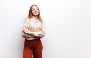 Genç güzel bir kadın omuz silkiyor, kafası karışık ve kararsız, kolları çapraz ve şaşkın bir şekilde beyaz duvara bakıyor.