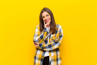 Genç ve güzel bir kadın mutlu görünüyor ve gülümsüyor. Elleri çenesinde, merak ediyor ya da soru soruyor. Turuncu duvarla seçenekleri karşılaştırıyor.