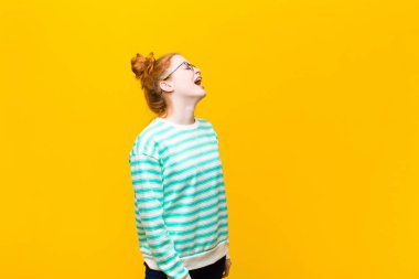 Genç kızıl saçlı kadın öfkeyle bağırıyor, agresif bağırıyor, stresli ve kızgın görünüyor turuncu duvara karşı.