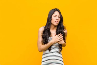 Genç ve güzel Latin kadın üzgün, yaralı ve kalbi kırık görünüyor, iki elini de kalbe yakın tutuyor, ağlıyor ve turuncu duvara karşı depresyona giriyor.