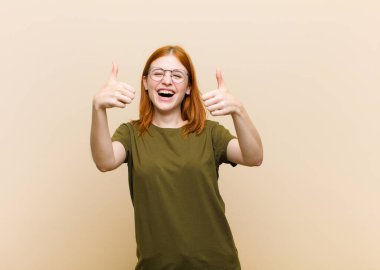 Genç kızıl saçlı güzel kadın gülümsüyor mutlu, pozitif, kendinden emin ve başarılı, iki başparmağı da havada.