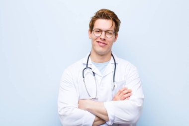 Genç kızıl saçlı doktor mutlu, gururlu ve memnun görünüyor. Kollarını mavi duvara dolamış gülümsüyor.