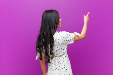 Fotokopi alanında duran ve objeye işaret eden genç Latin kadın, mor duvara karşı dikiz aynası.