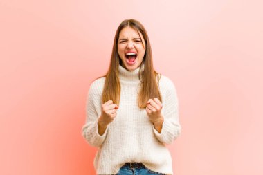 Genç ve güzel bir kadın sinirli sinirli, sinirli bakışlar ve yumruklarla pembe arka plana karşı öfkeli bir şekilde bağırıyor.