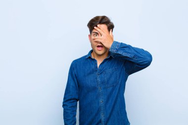 Genç yakışıklı adam şok olmuş, korkmuş ya da korkmuş görünüyor. Yüzü elle kaplanmış ve parmaklarının arasından mavi arka plana bakıyor.