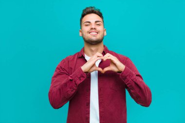 Genç İspanyol adam gülümsüyor ve mutlu, sevimli, romantik ve aşık oluyor. Mavi arka planda iki eliyle kalbi şekillendiriyor.
