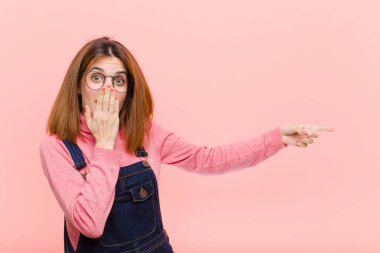 Genç, güzel bir kadın kendini mutlu, şok olmuş ve şaşırmış hissediyor. Ağzını eliyle kapatıyor ve pembe arka plana karşı yanal fotokopi alanını işaret ediyor.