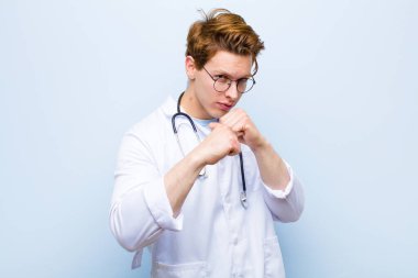 Genç kızıl saçlı doktor kendinden emin, sinirli, güçlü ve agresif görünüyor. Yumrukları mavi duvara karşı boks pozisyonunda dövüşmeye hazır.