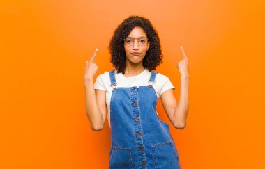 Kötü tavırlı, genç, siyah bir kadın gururlu ve agresif görünüyor, yukarı bakıyor ya da turuncu duvara yaslanmış ellerle dalga geçiyor.