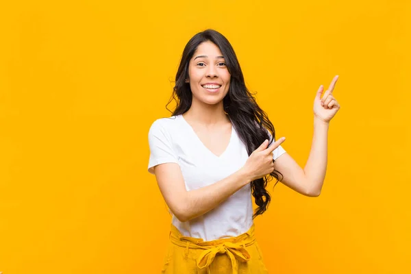 若いラテン系の女性は喜んで笑顔でオレンジの壁に対してコピースペースにオブジェクトを示す両方の手で上下を指しています — ストック写真