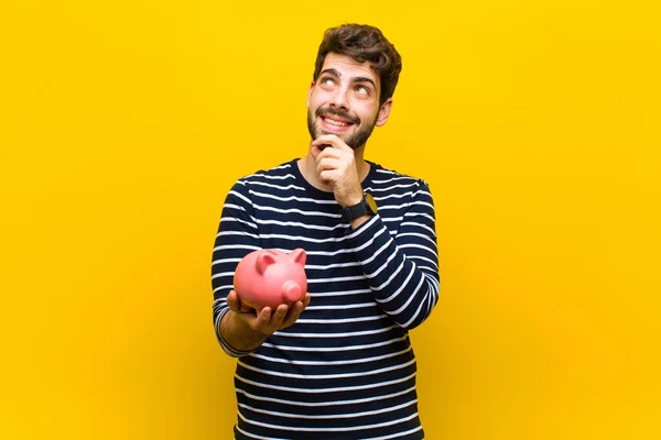 Jovem bonito homem segurando um porquinho banco contra laranja backgroun — Fotografia de Stock