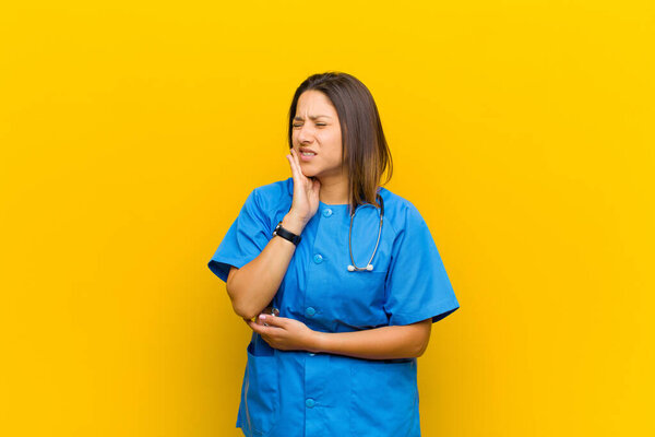 holding cheek and suffering painful toothache, feeling ill, miserable and unhappy, looking for a dentist isolated against yellow wall