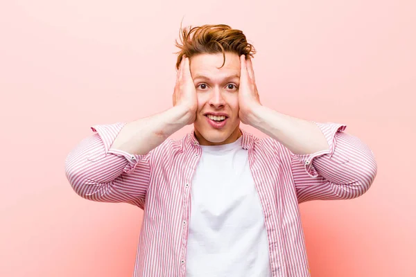 young red head man raising hands to head, open-mouthed, feeling extremely lucky, surprised, excited and happy against pink wall