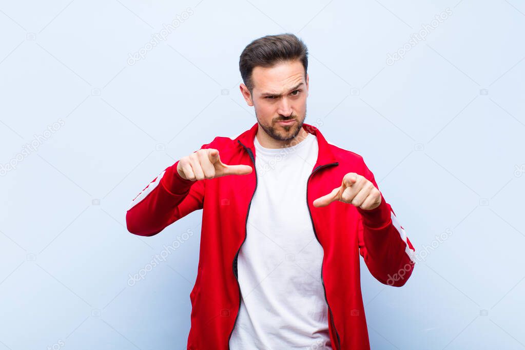 young handsome sports man or monitor pointing forward at camera with both fingers and angry expression, telling you to do your duty against flat wall
