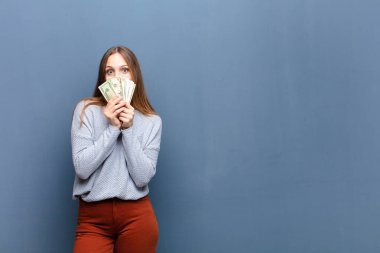 Bir dolar banknotuyla mavi duvara karşı duran genç ve güzel bir kadın.