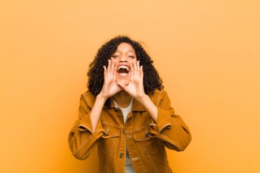 Genç, güzel siyah kadın mutlu, heyecanlı ve pozitif hissediyor. Elleri ağızda, turuncu duvara doğru bağırıyor.