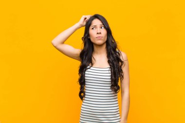 Genç, güzel, Latin bir kadın şaşkın ve kafası karışmış hissediyor. Kafasını kaşıyor ve turuncu duvara bakıyor.