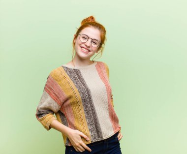 Genç, güzel, kızıl saçlı bir kadın yeşil duvara karşı neşeli, mutlu, kendinden emin ve rahat bir ifadeyle gülümsüyor.