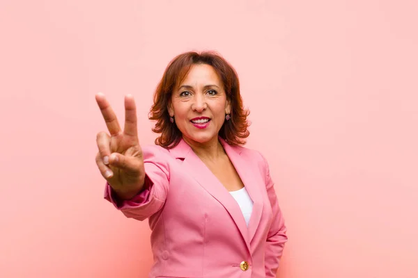 中年の女性は笑顔で幸せそして気楽でポジティブなピンクの壁に対して片手で勝利や平和をジェスチャー — ストック写真
