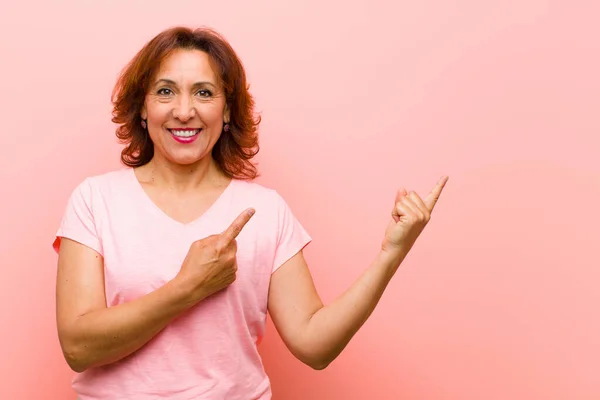middle age woman smiling happily and pointing to side and upwards with both hands showing object in copy space against pink wall