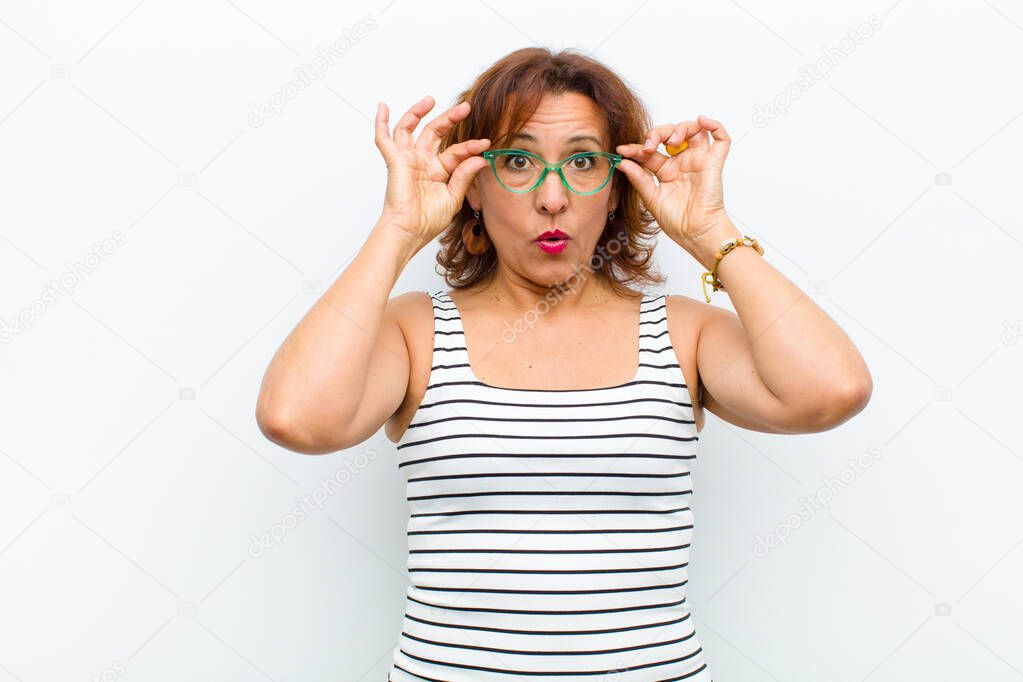 middle age pretty woman feeling shocked, amazed and surprised, holding glasses with astonished, disbelieving look against white wall
