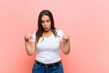 Genç, güzel, İspanyol bir kadın ağzı açık, iki eliyle aşağıya bakıyor, şaşkın, şaşkın ve pembe duvara karşı şaşırmış görünüyor.