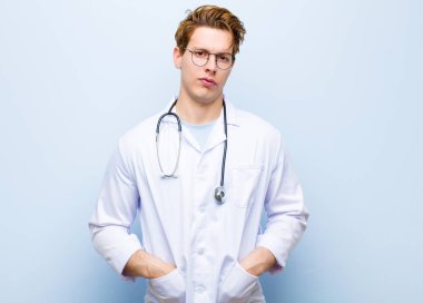 Genç kızıl saçlı doktor mavi duvara karşı neşeli, mutlu, kendinden emin ve rahat bir ifadeyle gülümsüyor.