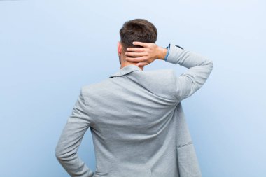 Genç işadamı düşünüyor ya da şüphe ediyor, kafasını kaşıyor, şaşkın ve şaşkın hissediyor, mavi arka planda ya da arka planda