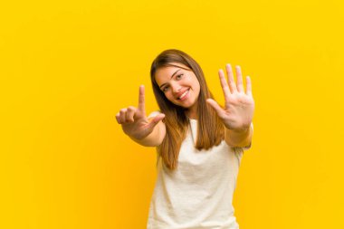 Genç, güzel bir bayan gülümsüyor ve dost canlısı görünüyor. Elleri önde, turuncu arka planda geriye doğru sayarak yedi ya da yedi numarayı gösteriyor.