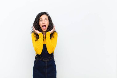 Genç ve güzel bir kadın beyaz duvara karşı beklenmedik bir sürprizden dolayı şaşkın, heyecanlı, gülen, şaşkın ve mutlu hissediyor.