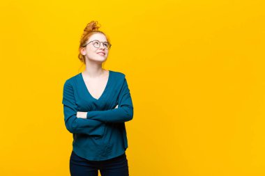 Genç kızıl saçlı kadın kendini mutlu, gururlu ve umutlu hissediyor, merak ediyor ya da düşünüyor, kollarını turuncu duvara dayamış çapraz kollarıyla uzayın fotokopisini çekmek istiyor.