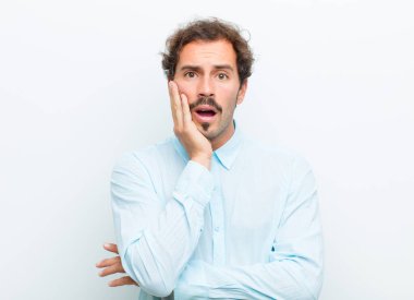 Genç yakışıklı adam şok ve inançsızlık içinde, elleri yanak ve kolları çaprazlanmış, sersemlemiş ve beyaz duvara karşı şaşkın hissediyordu.