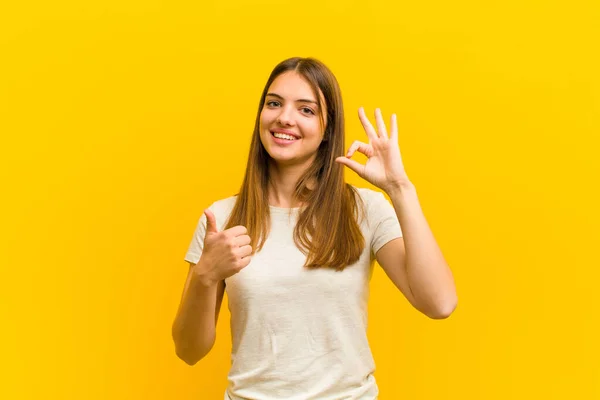 若い女性は幸せを感じ 満足し 大丈夫で親指を立ててジェスチャーを見せ オレンジの背景に微笑んだ — ストック写真