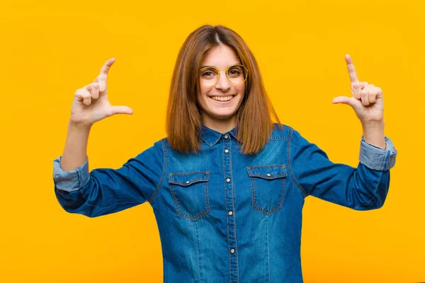 young pretty woman framing or outlining own smile with both hands, looking positive and happy, wellness concept against yellow background