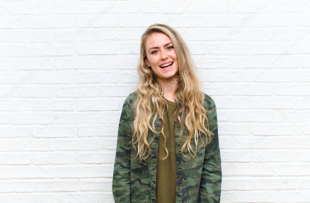 young blonde woman with a big, friendly, carefree smile, looking positive, relaxed and happy, chilling against brick wall background