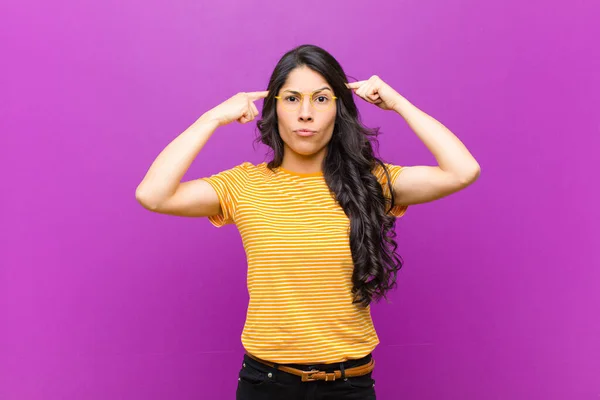 young pretty latin woman with a serious and concentrated look, brainstorming and thinking about a challenging problem against purple wall