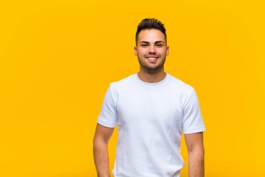Genç İspanyol adam mutlu bir şekilde gülümsüyor, eli kalçasında ve kendinden emin, pozitif, gururlu ve turuncu duvara karşı dost canlısı bir tavırla.