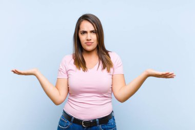 Genç, güzel bir kadın şaşkın, kafası karışmış ve stresli görünüyor. Farklı seçenekler arasında merak ediyor, mavi arka planda belirsizlik hissediyor.