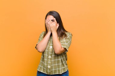 Genç, güzel bir kadın korkmuş ya da utanmış hissediyor, dikizliyor ya da gözleri yarı kapalı turuncu duvara yaslanmış bir şekilde dikizliyor.