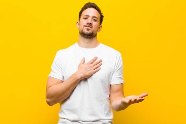 Genç yakışıklı adam mutlu ve aşık, bir eli kalbinin yanında gülümsüyor ve diğer eli turuncu duvara doğru uzanıyor.